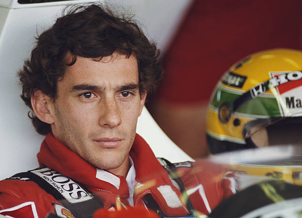 Ayrton Senna parado olhando dentro do cockpit, com o capacete ao lado
